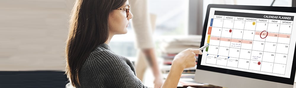 A woman wearing glasses refers to a calendar timeline on her computer.